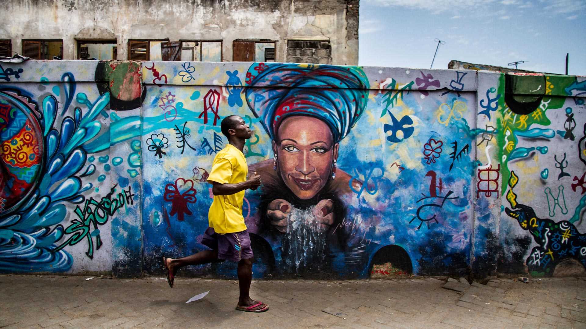 A man in Ghana running in the street in front of a wall painted with urban art